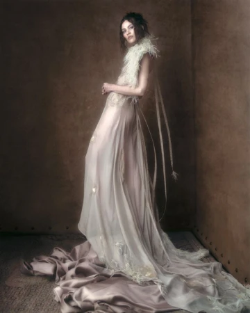Model in a flowing white gown with feathered neckline posing against a dark textured studio wall.