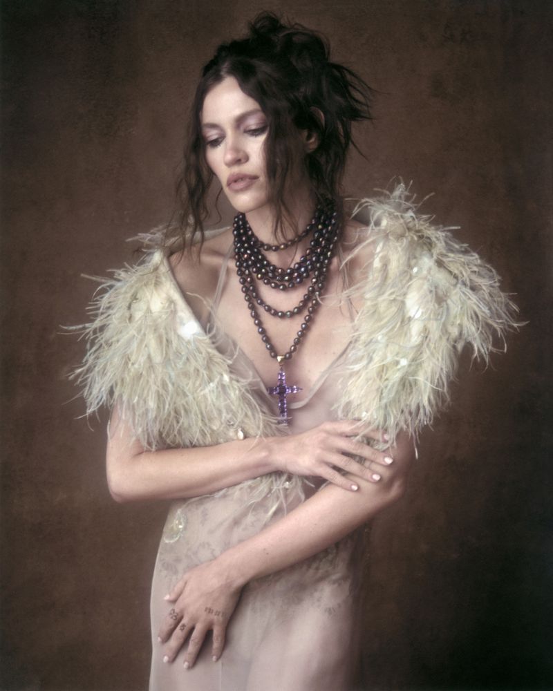 Model wearing feathered shoulders, layered pearl necklaces and a cross pendant against a brown textured background