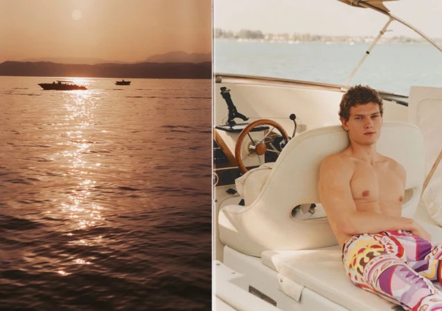 A shirtless male model wearing patterned trousers seated on a boat during sunset with calm sea views.
