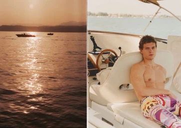 A shirtless male model wearing patterned trousers seated on a boat during sunset with calm sea views.