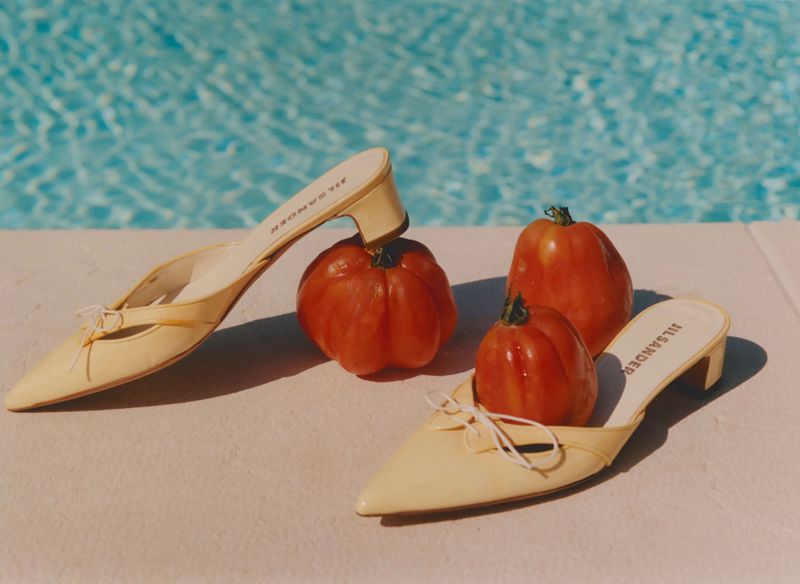Two pairs of yellow heeled mules arranged with ripe red tomatoes beside a shimmering blue swimming pool.
