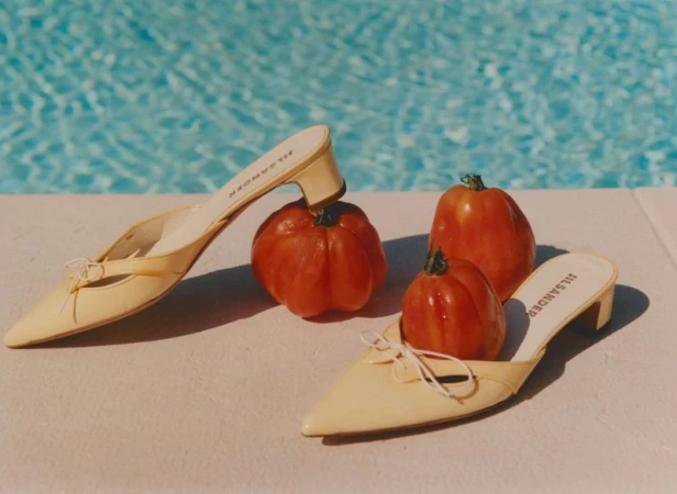 Two pairs of yellow heeled mules arranged with ripe red tomatoes beside a shimmering blue swimming pool.