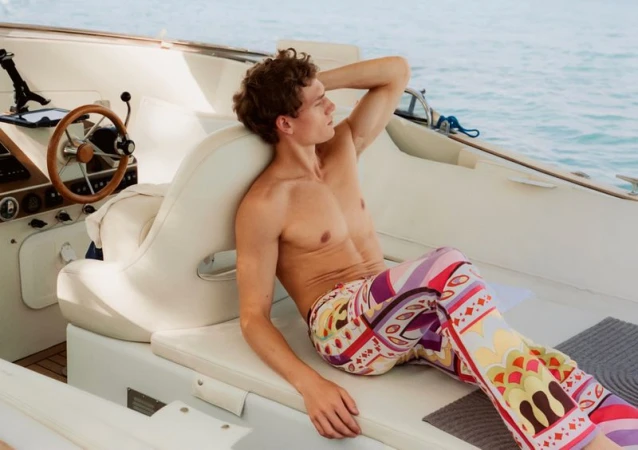 Shirtless man reclining on a boat deck wearing colorful patterned trousers against open water.