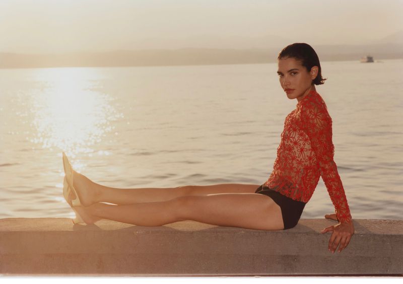 Model wearing a red lace top and black bottoms seated on a stone ledge by a body of water during sunset.