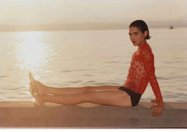 Model wearing a red lace top and black bottoms seated on a stone ledge by a body of water during sunset.