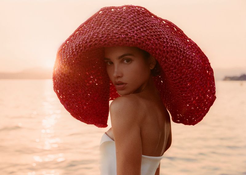Model wearing a large red woven sun hat and white top looking back over shoulder against a sunset ocean background