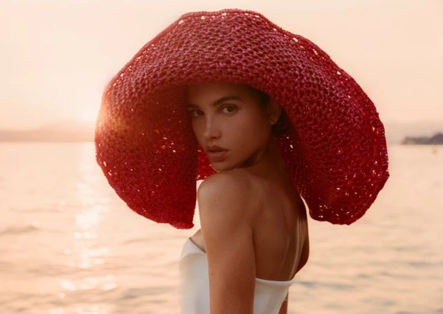 Model wearing a large red woven sun hat and white top looking back over shoulder against a sunset ocean background