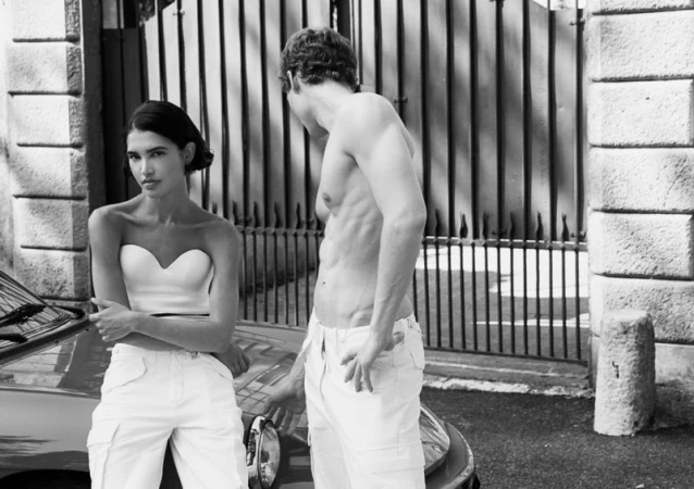 Black and white editorial fashion image featuring two models posing near a car in front of a stone building exterior.