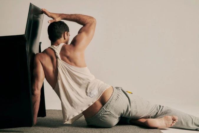 Athletic man wearing a white tank top and grey sweatpants seated on the floor leaning against a black chair.