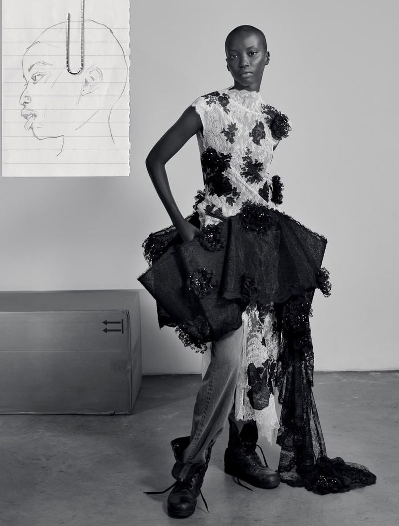 Black and white fashion portrait of a model wearing a lace dress and boots with a paper sketch pinned to the background.
