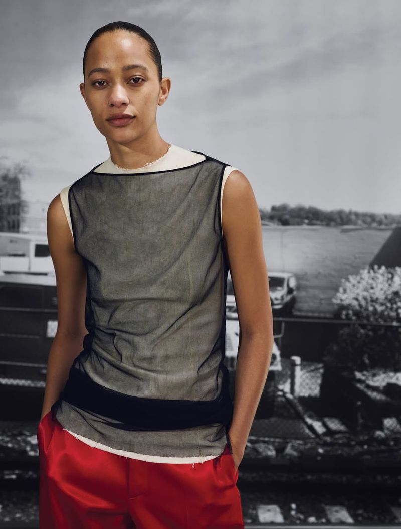 Model wearing a sheer sleeveless top and bright red trousers standing against a muted urban backdrop for British Vogue.