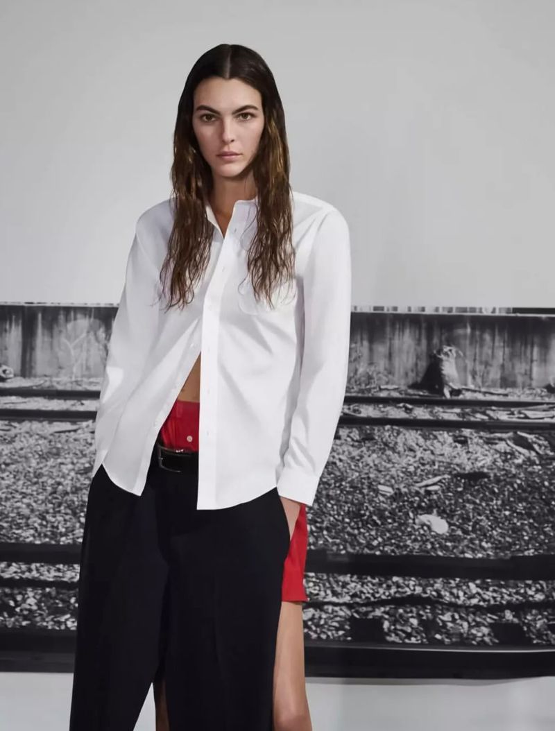 Model wearing a white button-down shirt and black trousers standing against a textured backdrop for British Vogue.