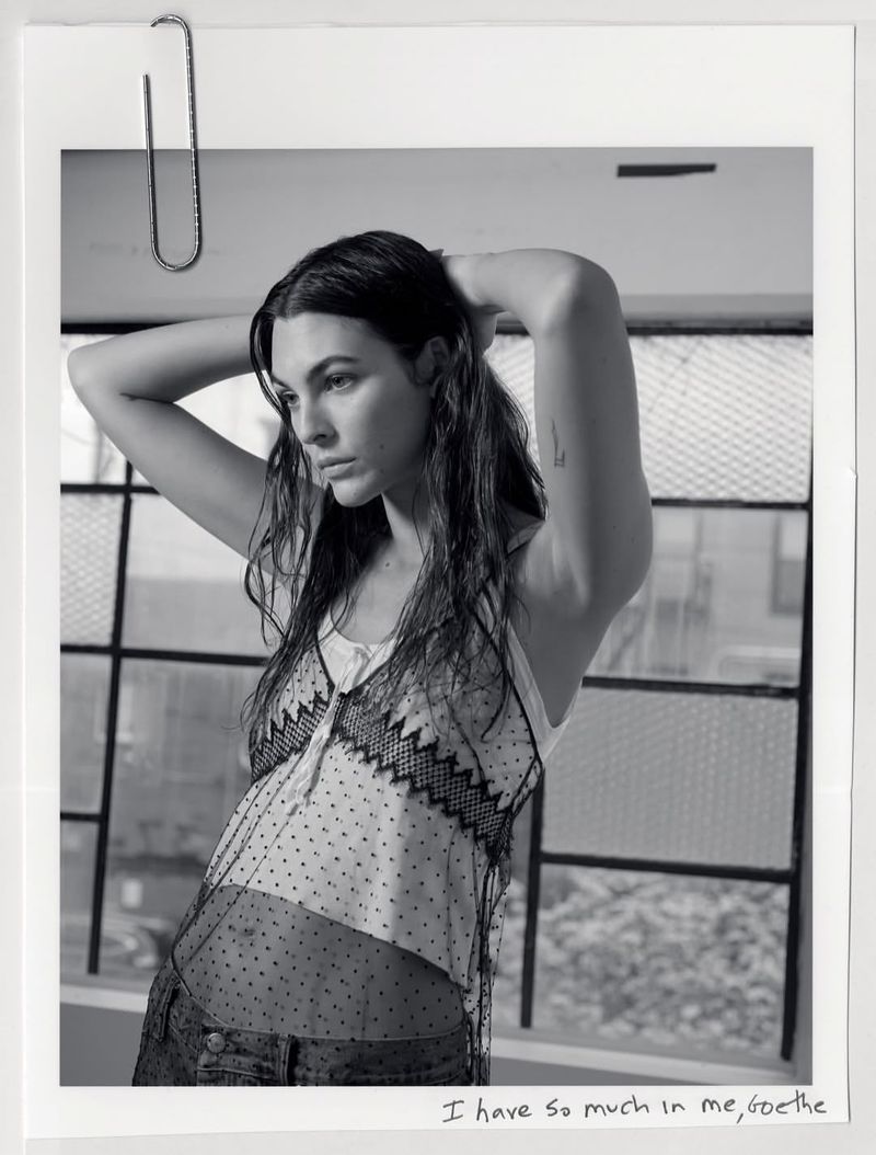 Black and white portrait of a woman with dark hair posing with hands behind her head against a window frame.