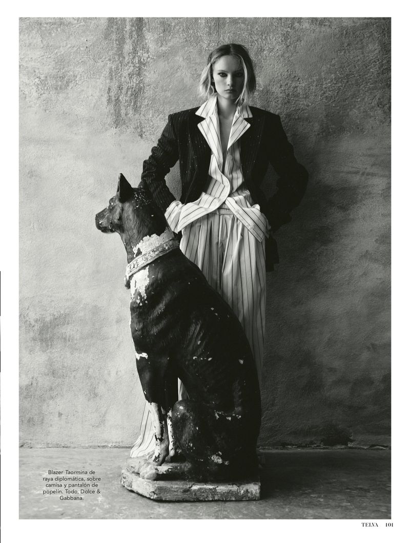 Model standing behind a large dog statue wearing a dark pinstripe blazer and matching trousers against a textured stone wall.