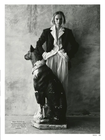 Model standing behind a large dog statue wearing a dark pinstripe blazer and matching trousers against a textured stone wall.