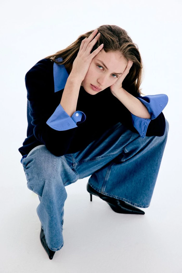 Model crouching with hands to head wearing black top with blue cuffs and wide-leg denim jeans against a white background