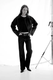 Model wearing dark jacket and trousers standing against a bright white background with studio lighting equipment visible on the right.