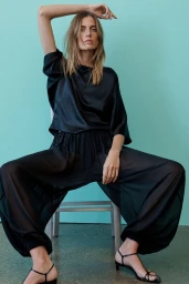 Model in black satin top and sheer trousers sitting on a chair against a blue background
