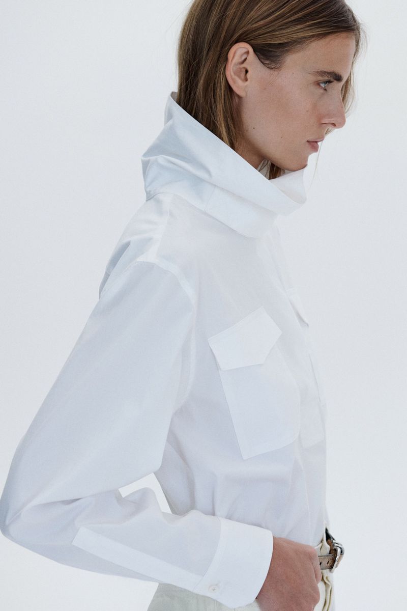 Profile view of a female model wearing a white utility shirt with a high collar against a plain white background
