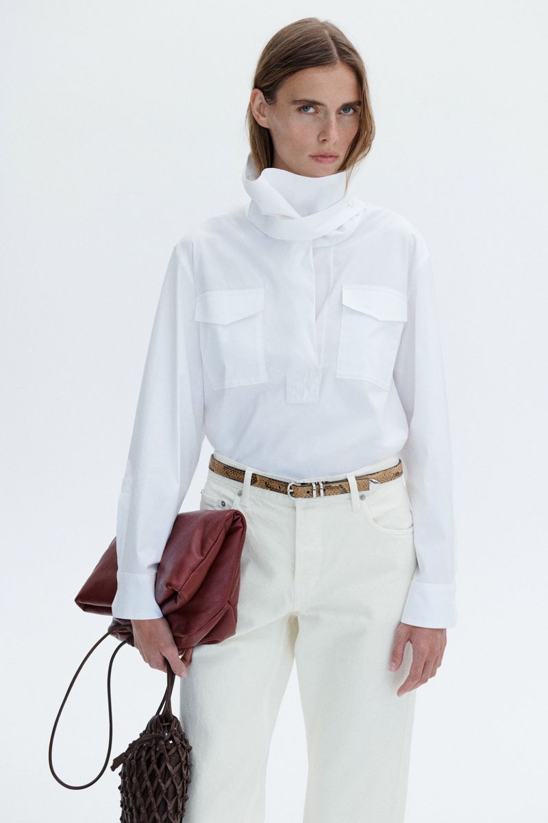 Model wearing a white blouse and trousers holding a dark red clutch against a solid white background
