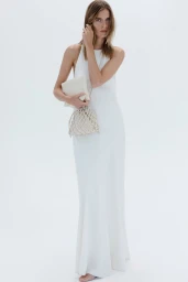 Model in white halter dress holding a cream pouch and woven net handbag against white