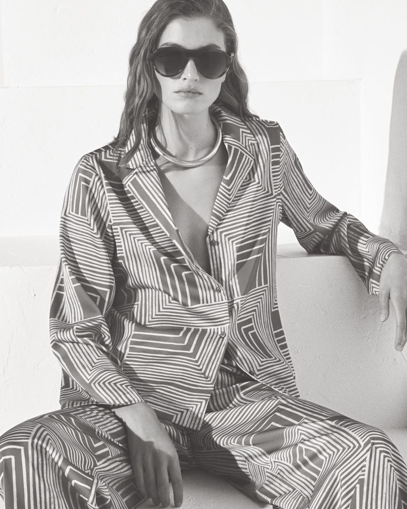 Model in patterned shirt and trousers wearing dark sunglasses against a white background