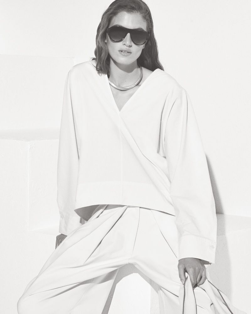 Model wearing dark sunglasses and a white V-neck top against a minimal white studio background.