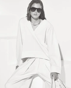 Model wearing dark sunglasses and a white V-neck top against a minimal white studio background.