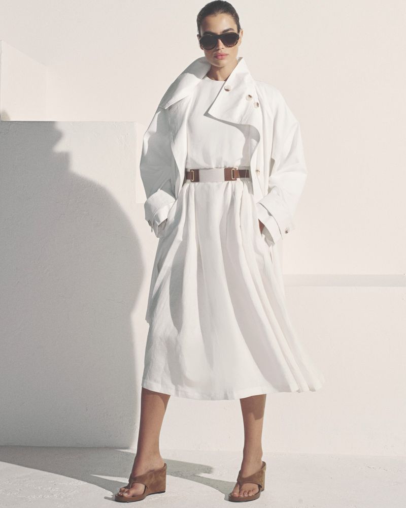 Model in dark sunglasses wearing an oversized white coat and dress belted with brown leather against a light background
