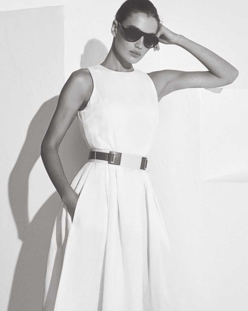 Model wearing dark sunglasses and a belted white dress posing against a plain wall in a monochrome lookbook shot.