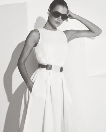 Model wearing dark sunglasses and a belted white dress posing against a plain wall in a monochrome lookbook shot.