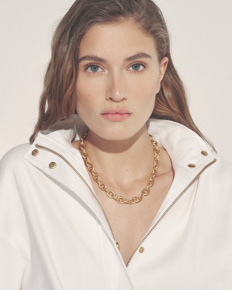 Model wearing a white jacket and gold chain necklace against a seamless background for Donna Karan