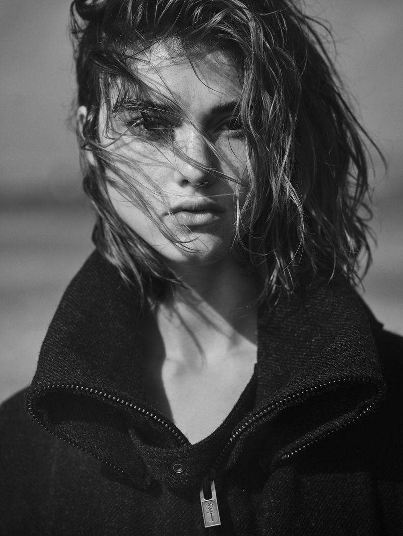 Monochrome portrait of a woman with wet hair, wearing a dark wool coat against a blurred grey background. Focus on face.
