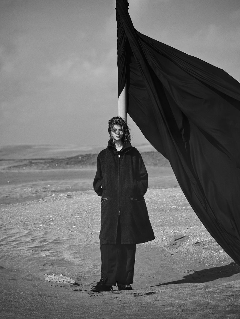 An adult female stands on a sandy surface near a draped black cloth set against a backdrop of dunes and a blue sky in black and white.