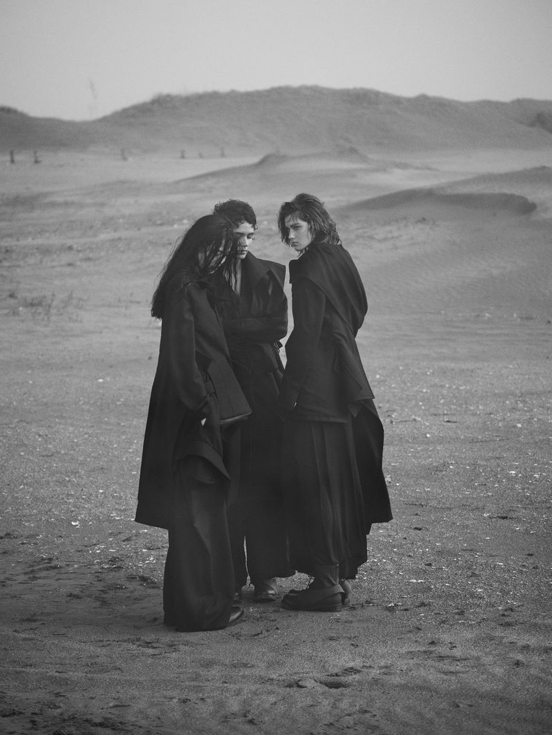 Three people in dark, flowing coats gather in a bleak, grainy landscape under an overcast sky in this black and white fashion photograph.