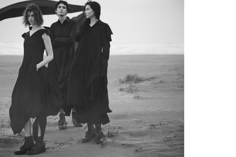 Three women in dark, flowing dresses and combat boots pose in a desolate landscape, creating a high-fashion, editorial look.