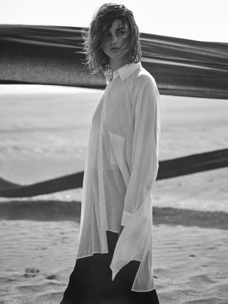A woman in a white shirt poses on a sandy beach with a dark scarf waving in the background, black and white fashion editorial.