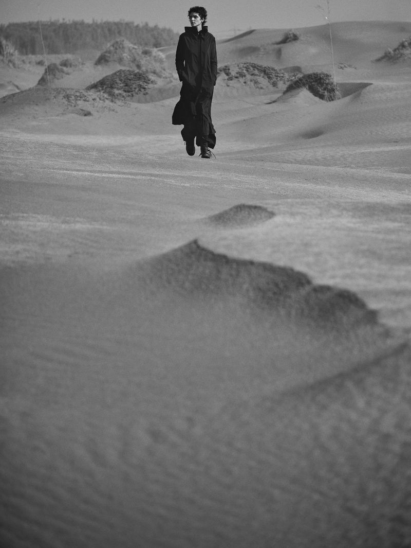 Monochrome portrait of a person in a long dark coat walking across sand dunes on a sunny day, creating a stark, windswept scene.