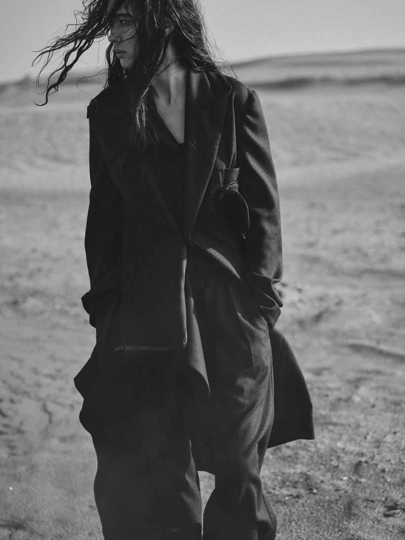 A woman in a dark outfit stands in a black and white outdoor fashion shot. The background is a sandy landscape, and her hair is windblown.