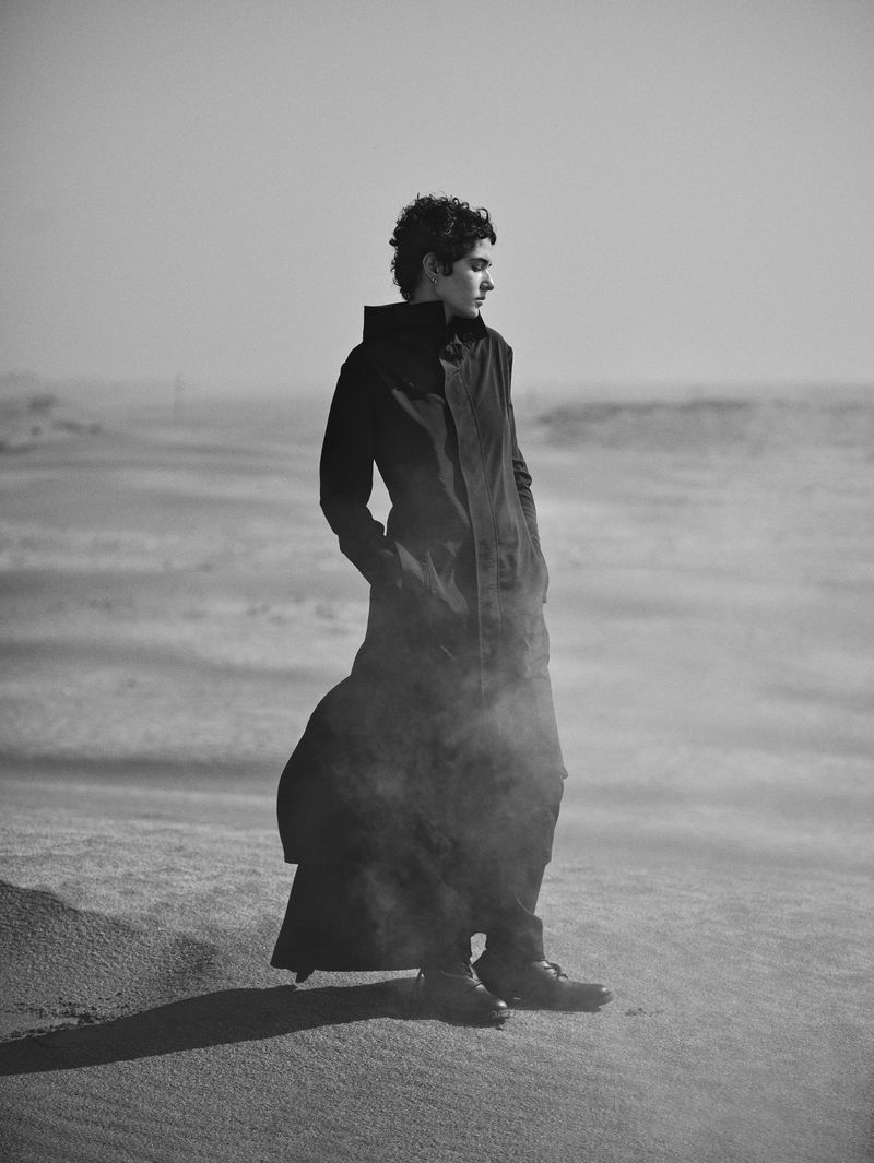 Monochromatic portrait of a person in a long dark coat standing in a sandy landscape, gazing off to the side with a neutral expression.