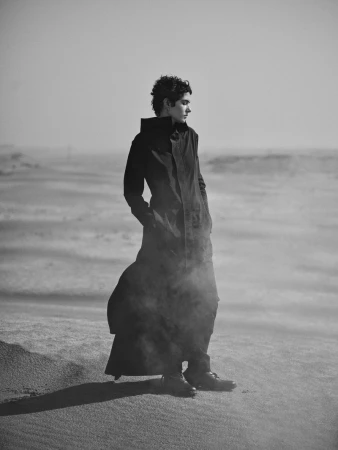 Monochromatic portrait of a person in a long dark coat standing in a sandy landscape, gazing off to the side with a neutral expression.