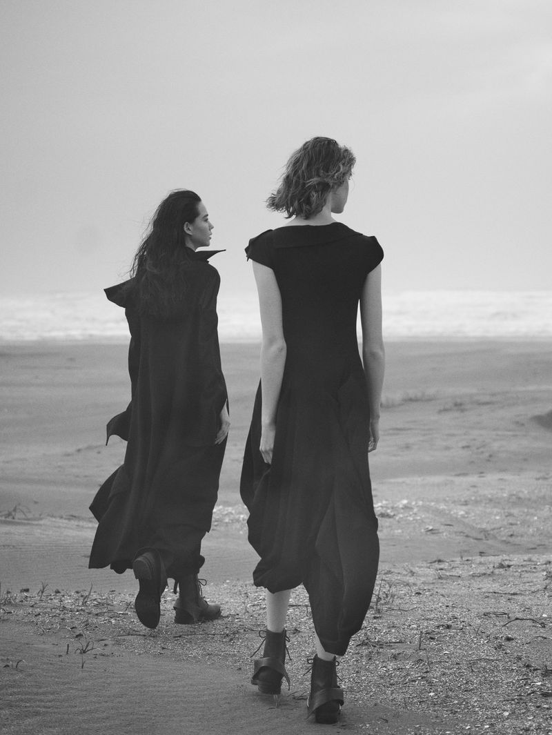Two women in dark clothing walk on a sandy beach under a cloudy sky. One wears a cape, the other a dress and boots, shot in black and white.