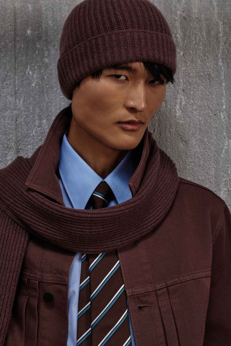 Model wearing a brown beanie, matching jacket, blue button-down shirt, and striped tie against a concrete wall