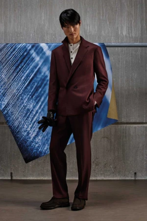 Model in a burgundy suit wearing black leather gloves posing against a textured background for Kiton KNT F/W 2026.