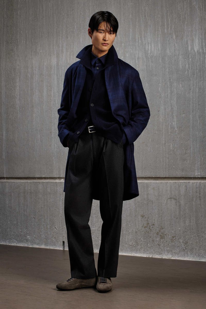 A man wearing a dark blue coat and trousers standing against a textured grey wall for the Kiton KNT F/W 2026 campaign.