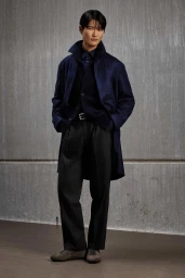 A man wearing a dark blue coat and trousers standing against a textured grey wall for the Kiton KNT F/W 2026 campaign.