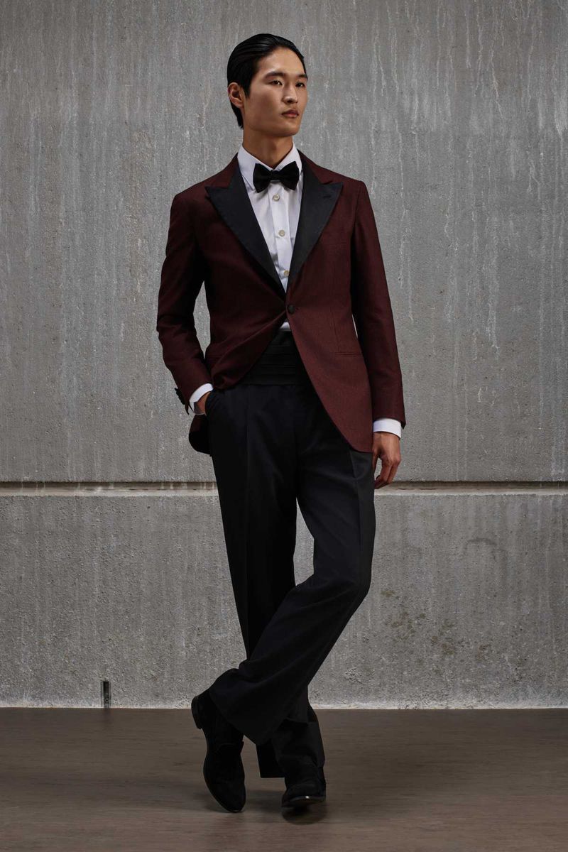 Model wearing a burgundy tuxedo jacket, black trousers, and bow tie standing against a grey concrete wall.