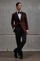 Model wearing a burgundy tuxedo jacket, black trousers, and bow tie standing against a grey concrete wall.