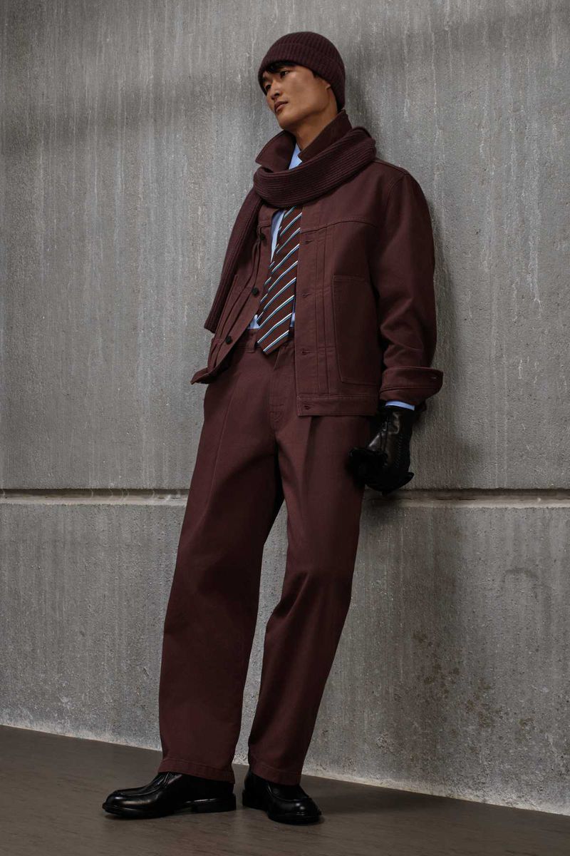Model leaning against a concrete wall wearing a maroon jacket, matching trousers, beanie, and a knitted scarf with a striped tie.