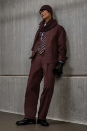 Model leaning against a concrete wall wearing a maroon jacket, matching trousers, beanie, and a knitted scarf with a striped tie.